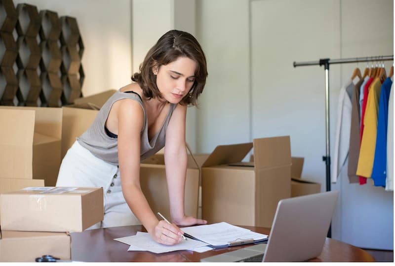 Boutique owner checking inventory paperwork surrounded by small batch shipment boxes and clothing rack