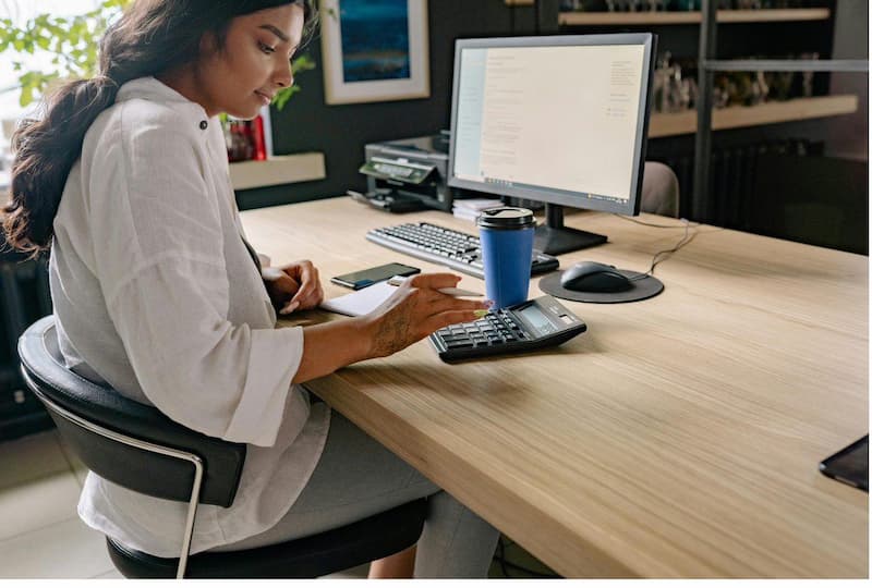 Small business owner calculating boutique inventory margins at her desk with a calculator and computer